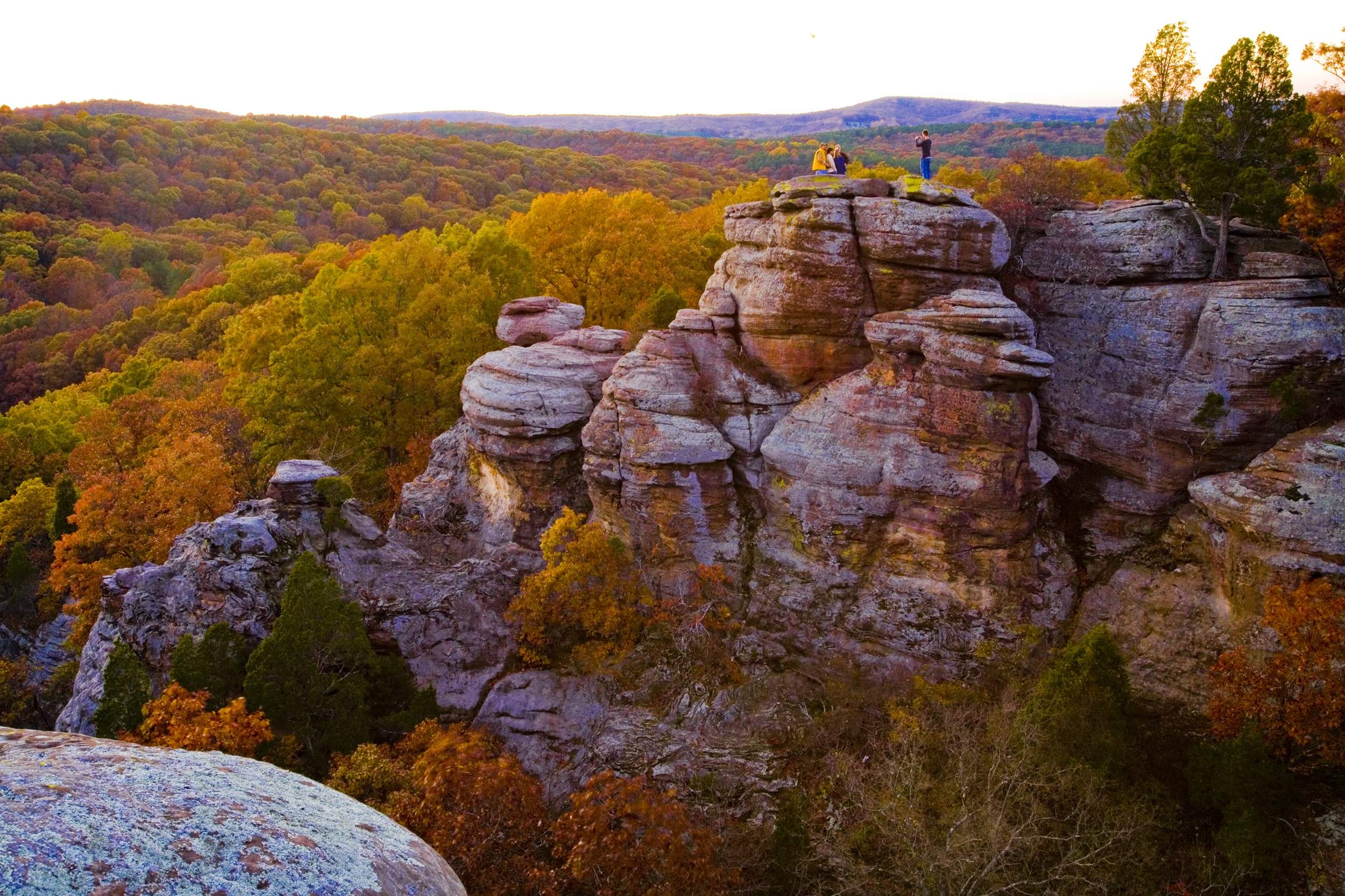 A distant group of four people posing for a photo atop rocky bluffs, overlooking forests of green and gold trees