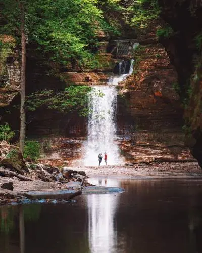 Two people standing in front of a waterfall, next to a serene pond.