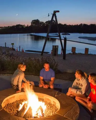 Four people sitting around a fire next to a lakeside beach at dusk